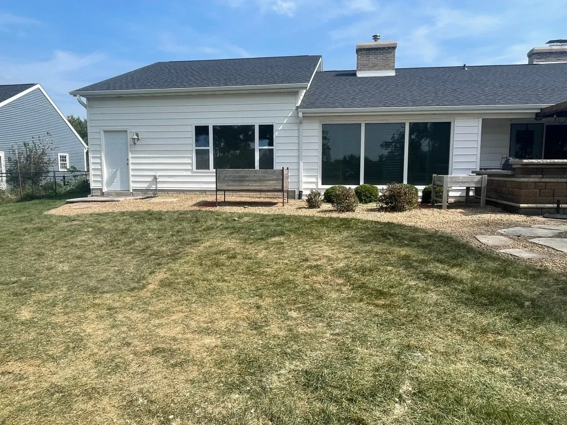 Backyard of a white house with dark windows and a black roof. A bench sits in front.