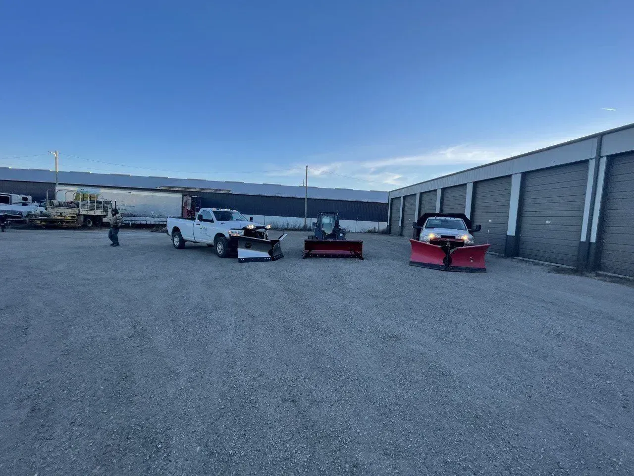 Three vehicles with plows parked outside storage units on a gravel lot under a blue sky.