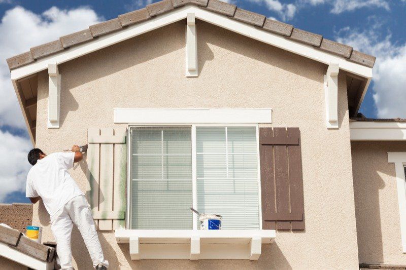 Painter in white overalls on a ladder painting shutters beige on a light-colored house.