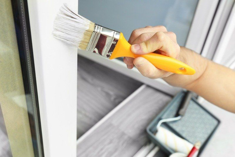 Hand with yellow paintbrush painting a white window frame. Gray roller and tray visible.