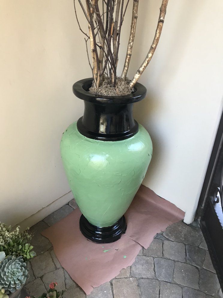 Large green vase with black top and base, containing dried branches, on a stone floor.