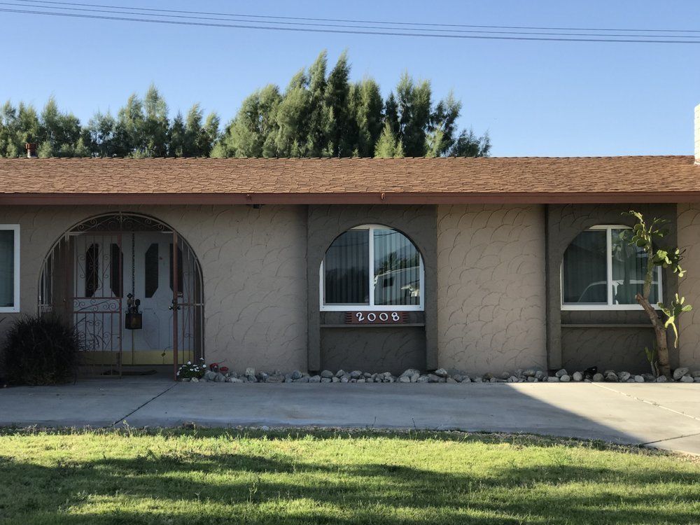 Tan single-story house with arched windows and brown roof, set against a backdrop of trees and a clear blue sky.