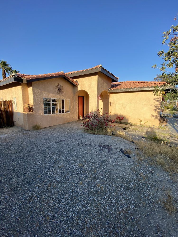 Tan stucco house with red tile roof, gravel driveway, and sparse landscaping under a blue sky.