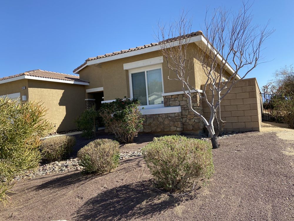 Tan house with bushes and a dry, bare tree, set against a clear blue sky.