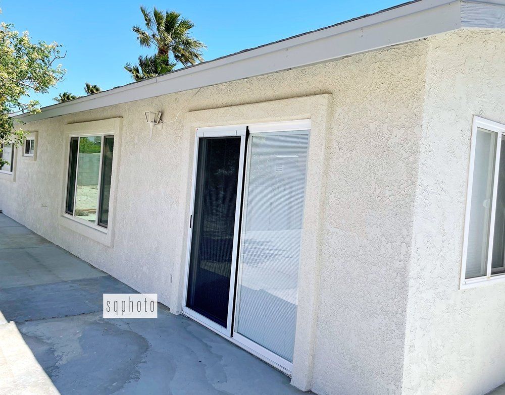 Exterior of a building with sliding glass door and windows, light stucco walls, and a blue sky.