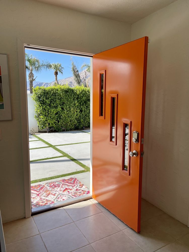 Open orange door revealing outdoor green hedge and palm trees.