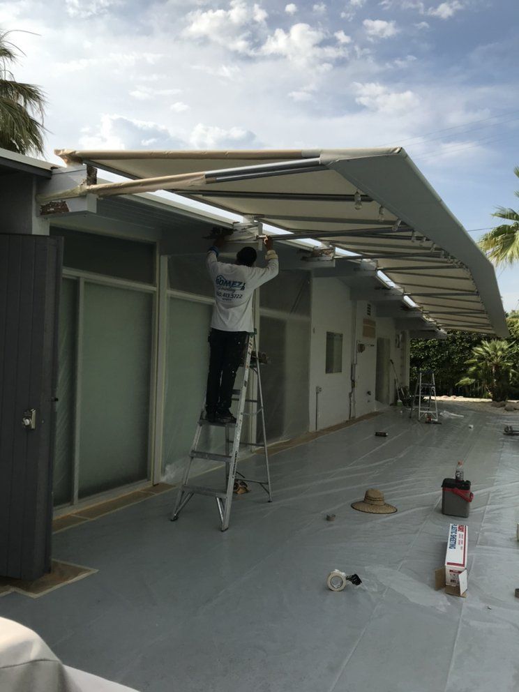 Man on ladder installing awning on a building exterior. Gray walls, blue sky, and a concrete patio are visible.