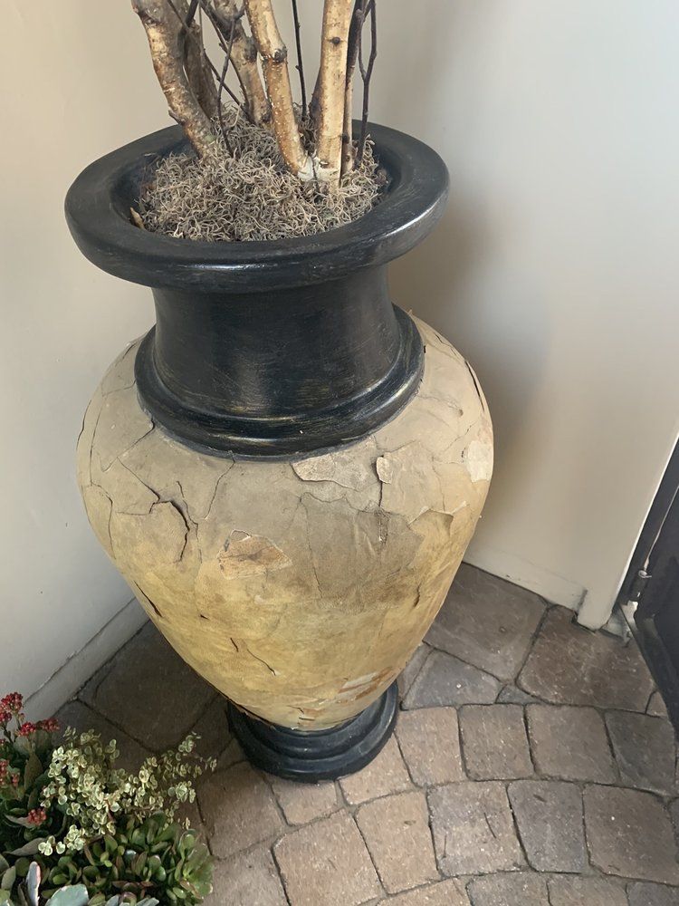 Large decorative urn with black trim and cracked tan finish, containing dried branches, on a stone floor.