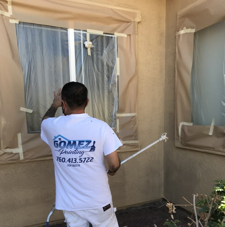 A person spraying paint on a house exterior. Windows are taped and covered.