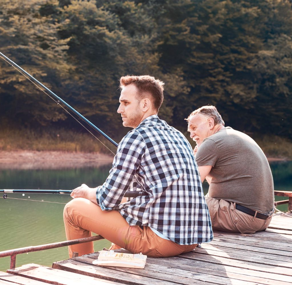 Two people fishing on a wooden dock overlooking a lake. One holds a fishing rod, the other watches.
