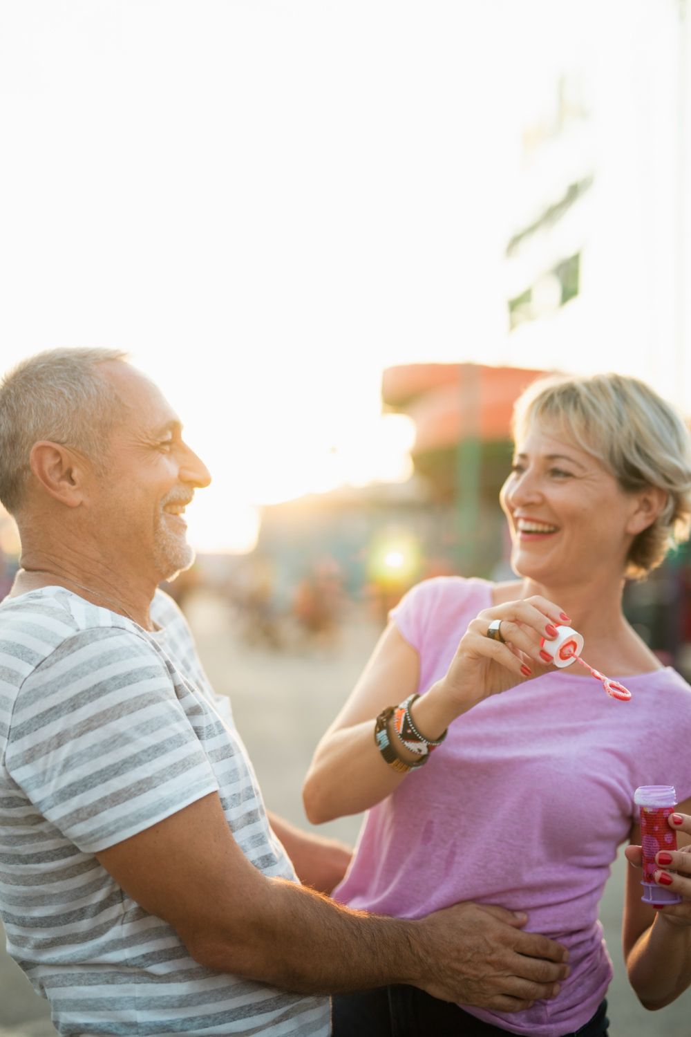 Couple laughs while blowing bubbles outdoors. Bright sunlight.