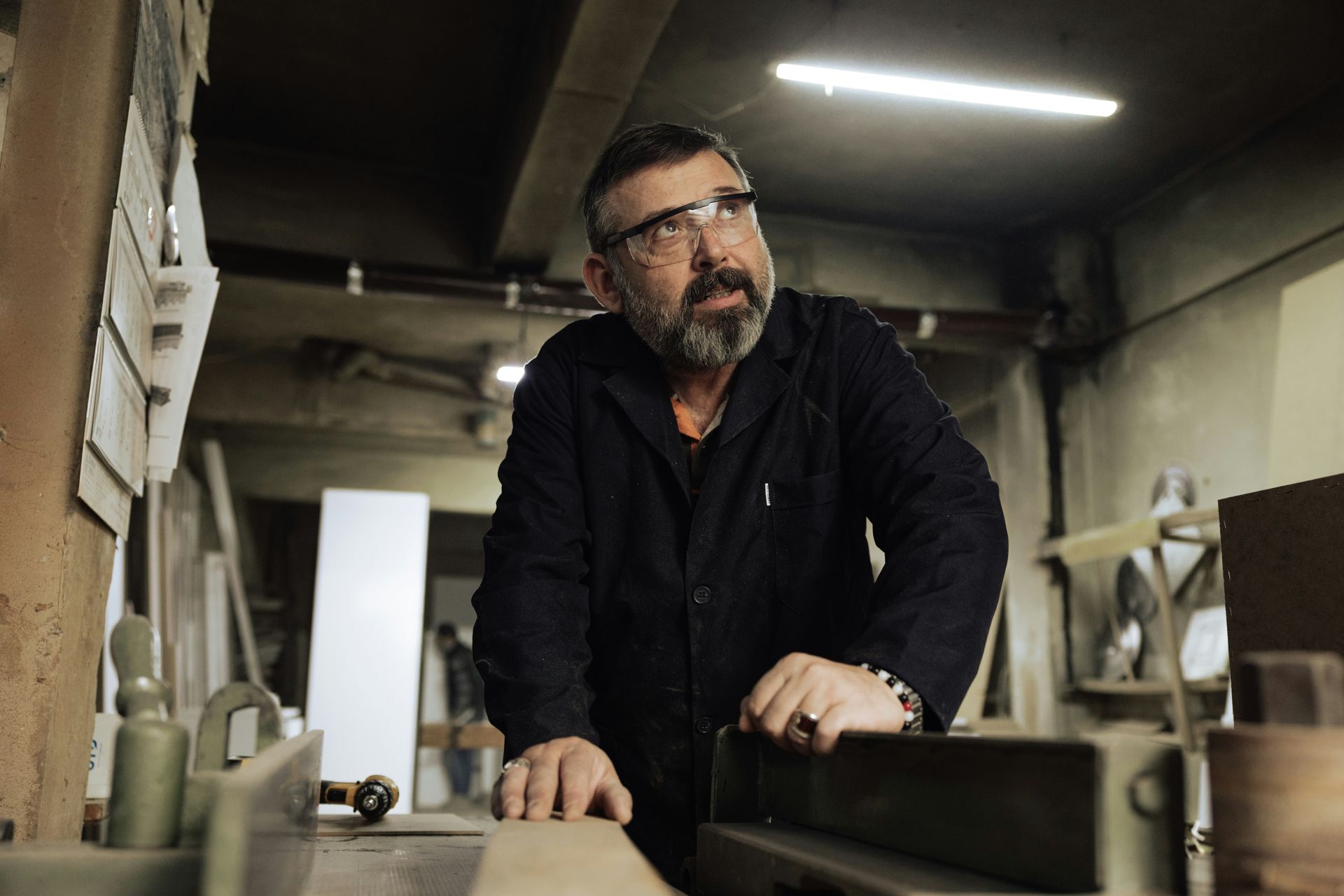 Man in a workshop looking upwards, wearing glasses. Dark clothing, wood and machinery in the background.