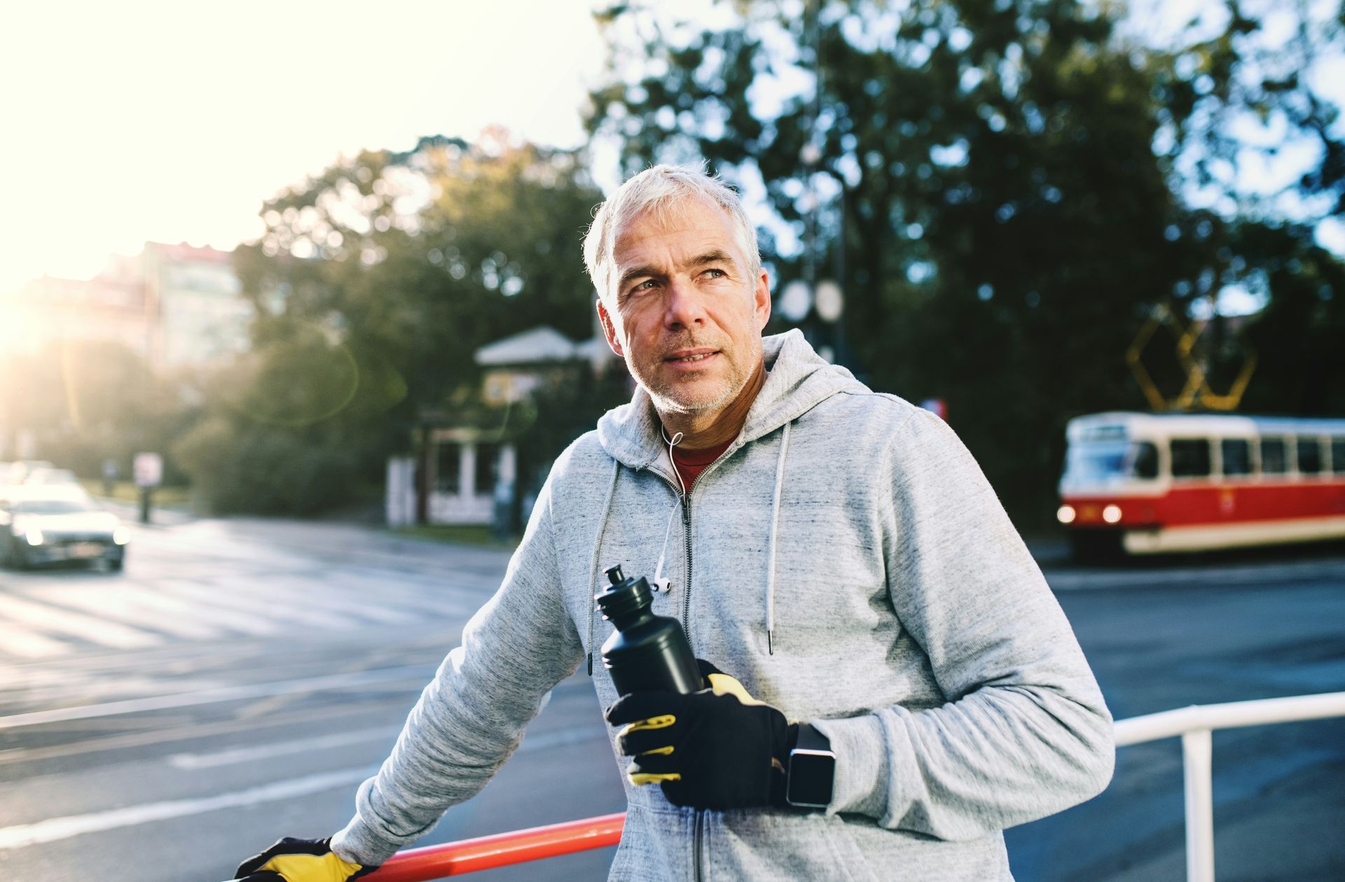 Man in gray hoodie and gloves holds water bottle near a street with a tram.