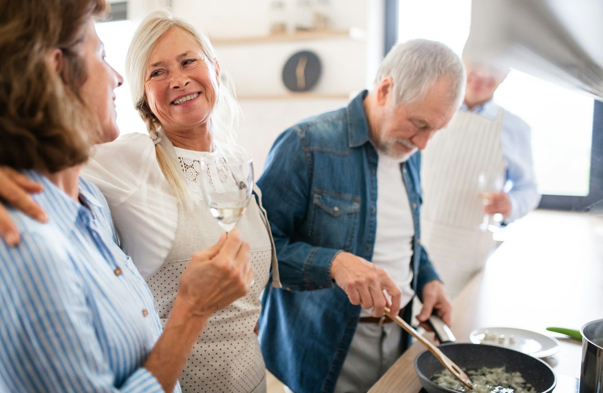 Group of older adults in a kitchen; one cooking, others smiling and holding drinks.