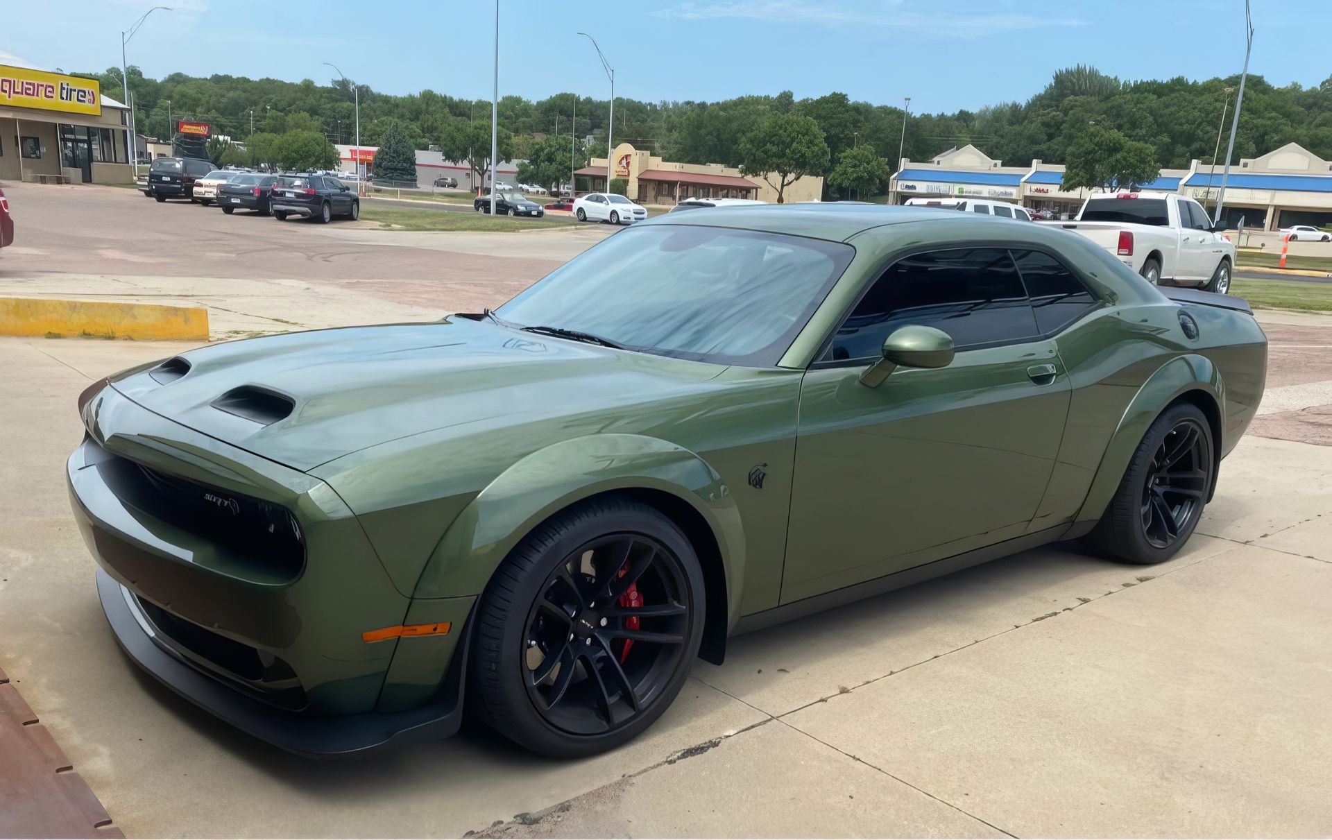 Matte olive green Dodge Challenger parked on a concrete lot under a cloudy sky.