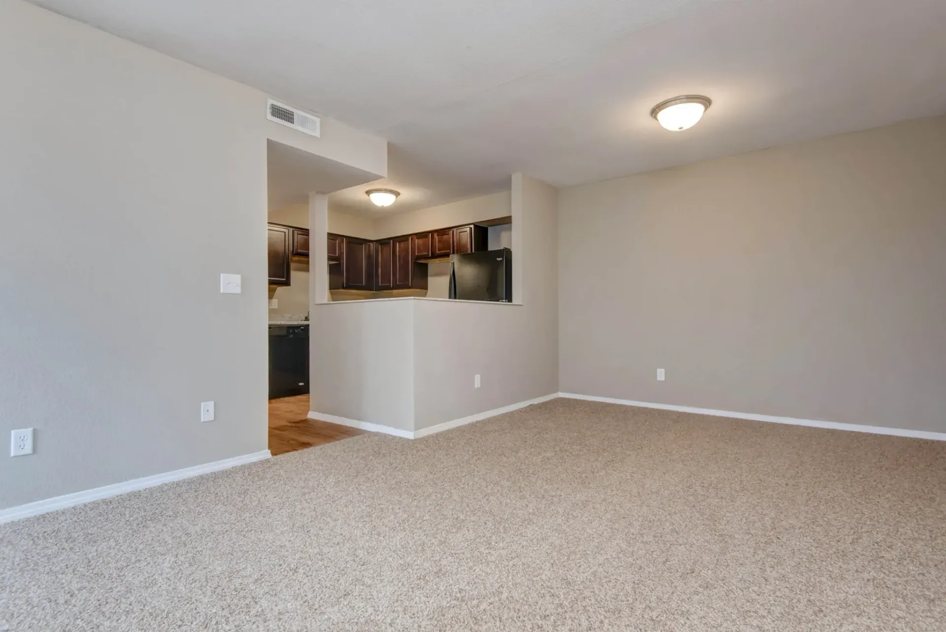 Photo of a carpeted living room with the kitchen in the background