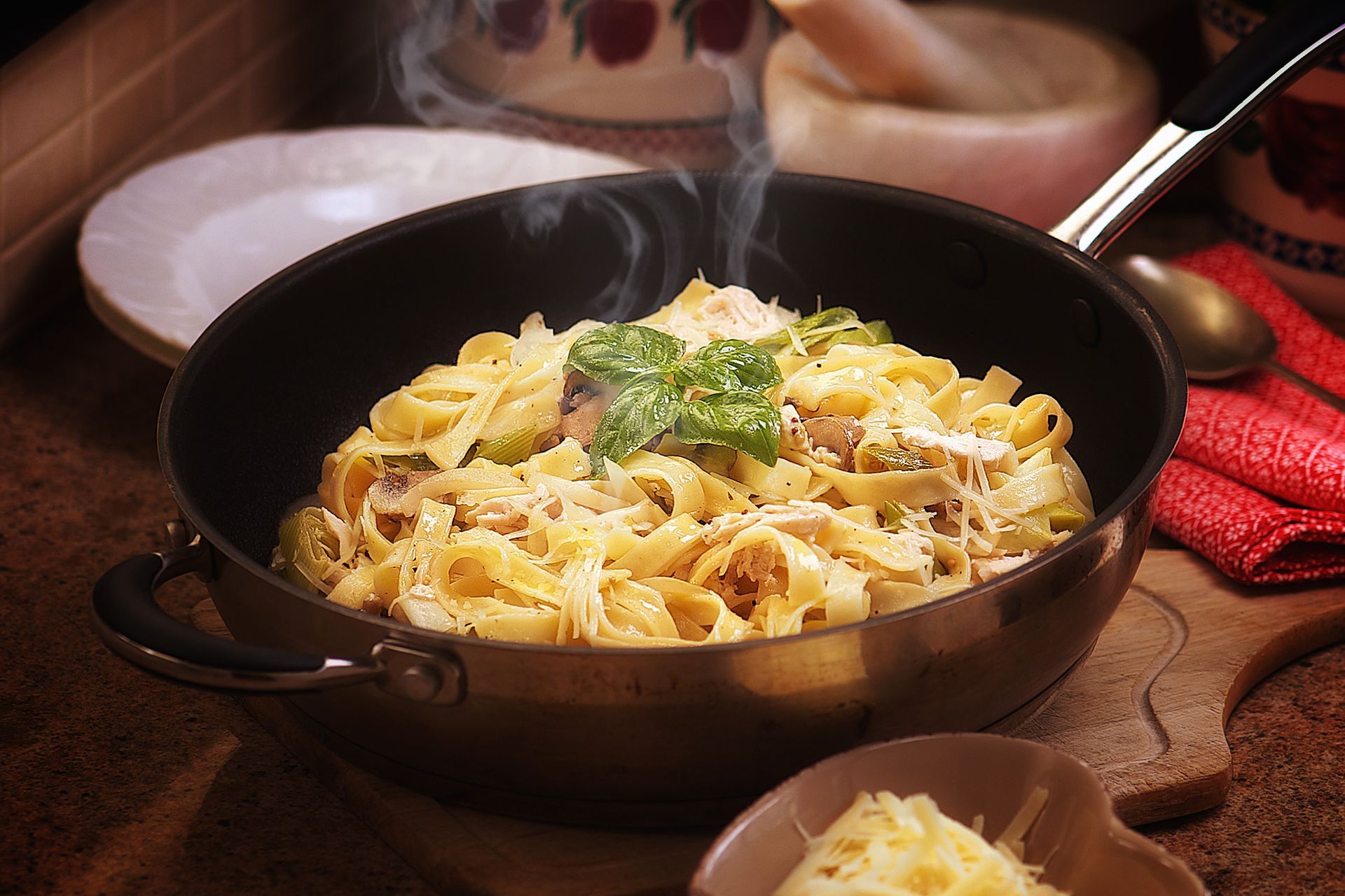 Steaming pasta dish in a pan, topped with cheese and basil. Wooden board and small bowl of cheese are visible.