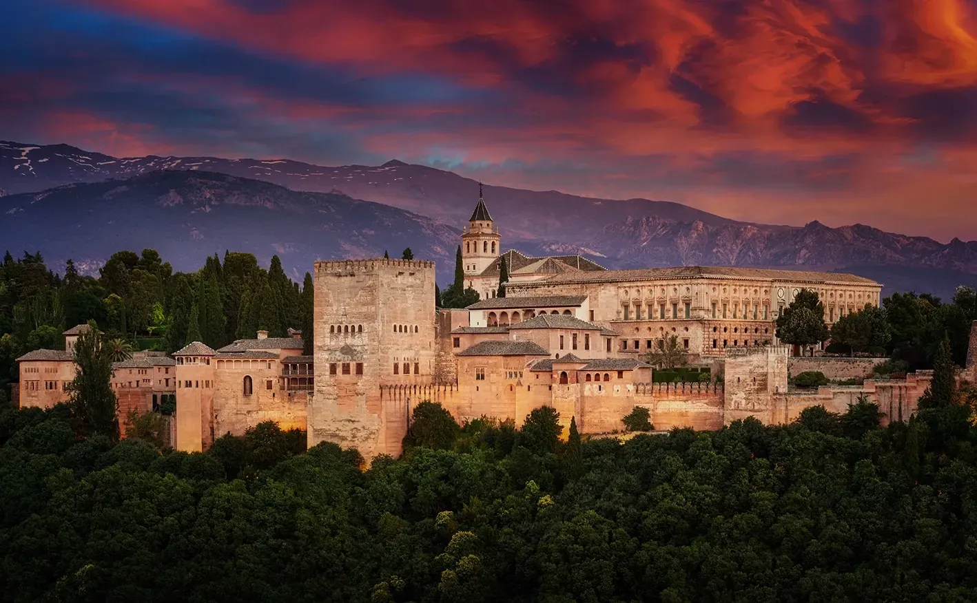 The Alhambra palace in Granada, Spain, lit against a sunset sky with a mountain backdrop.
