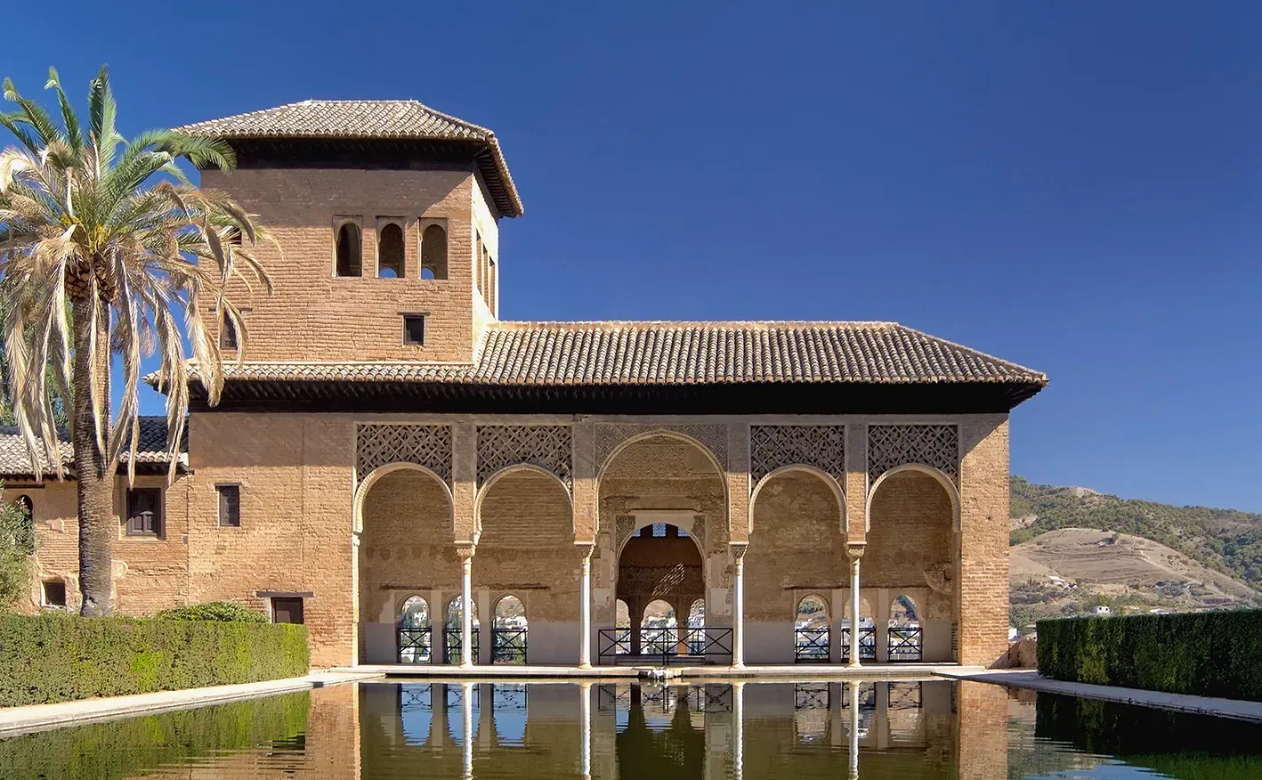Brick building with arches, reflecting in a pool. Tower, palm tree, and blue sky in background.