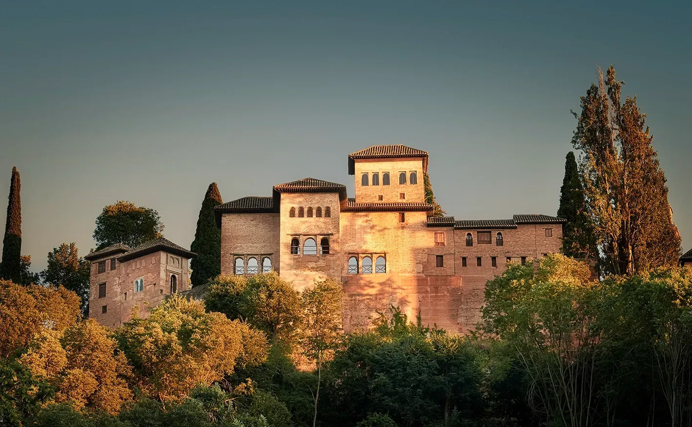 Granada's Alhambra Palace at sunset, bathed in warm light, set atop a hill, surrounded by trees.