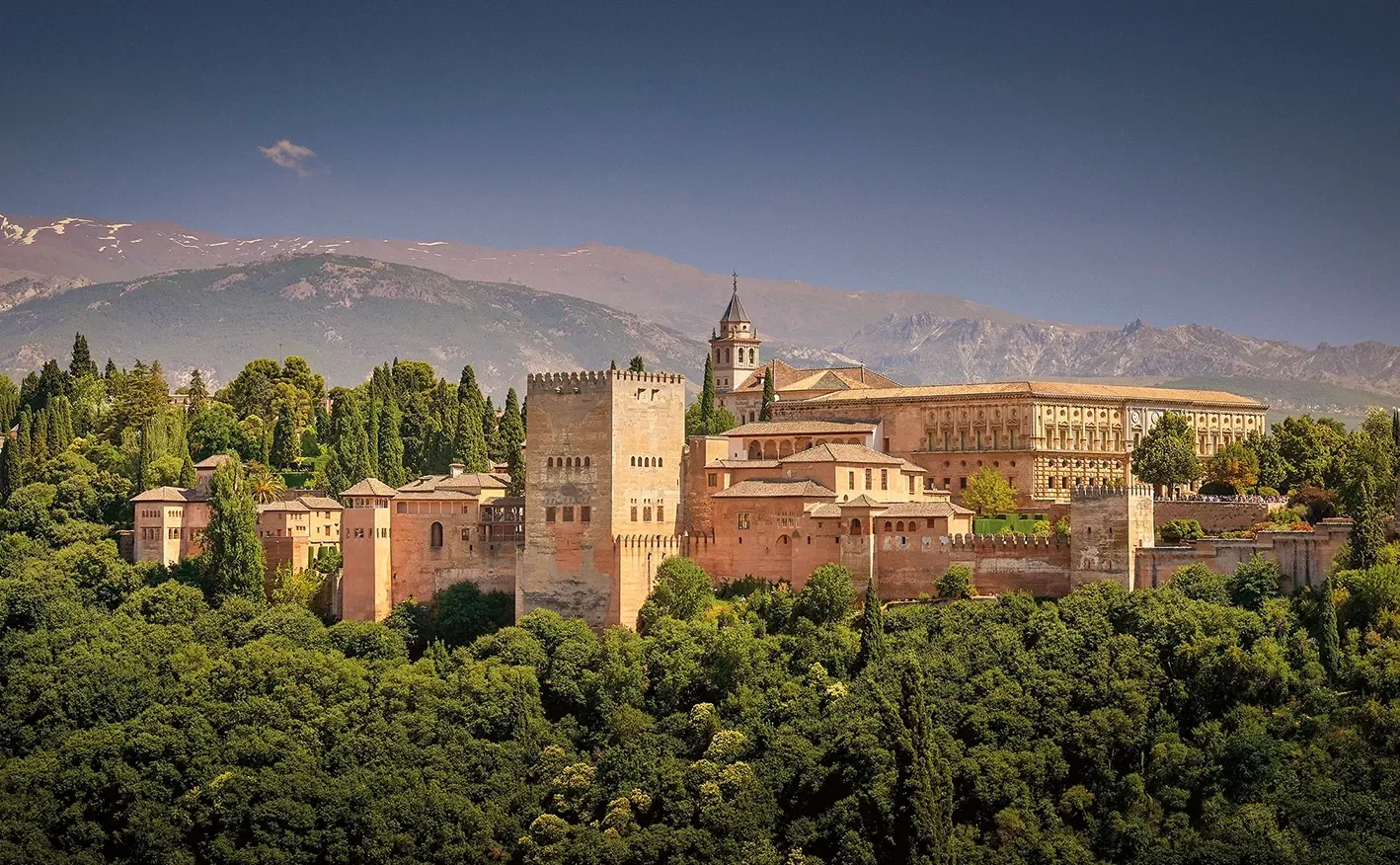 Moorish Alhambra palace in Granada, Spain, atop a green hill, with a backdrop of mountains and blue sky.