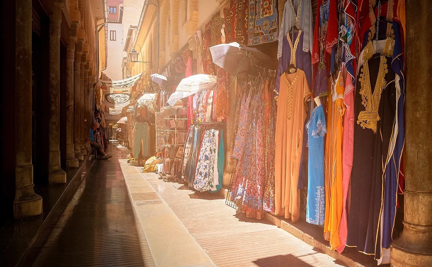 Narrow street with clothing displayed in shop windows; vibrant colors, sunlight.
