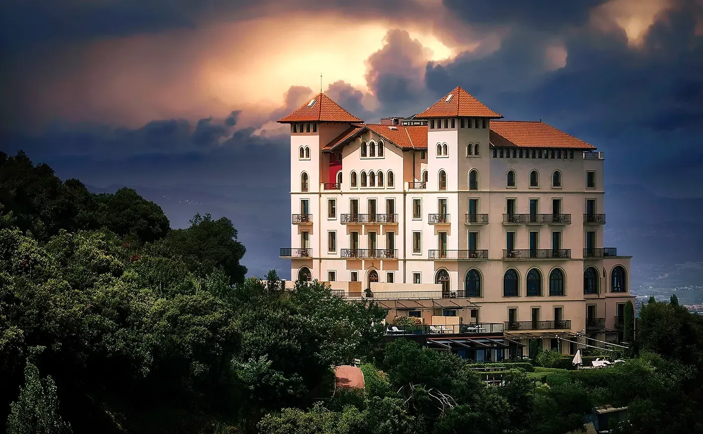White building with red-tiled roof on a hillside under a cloudy sky.