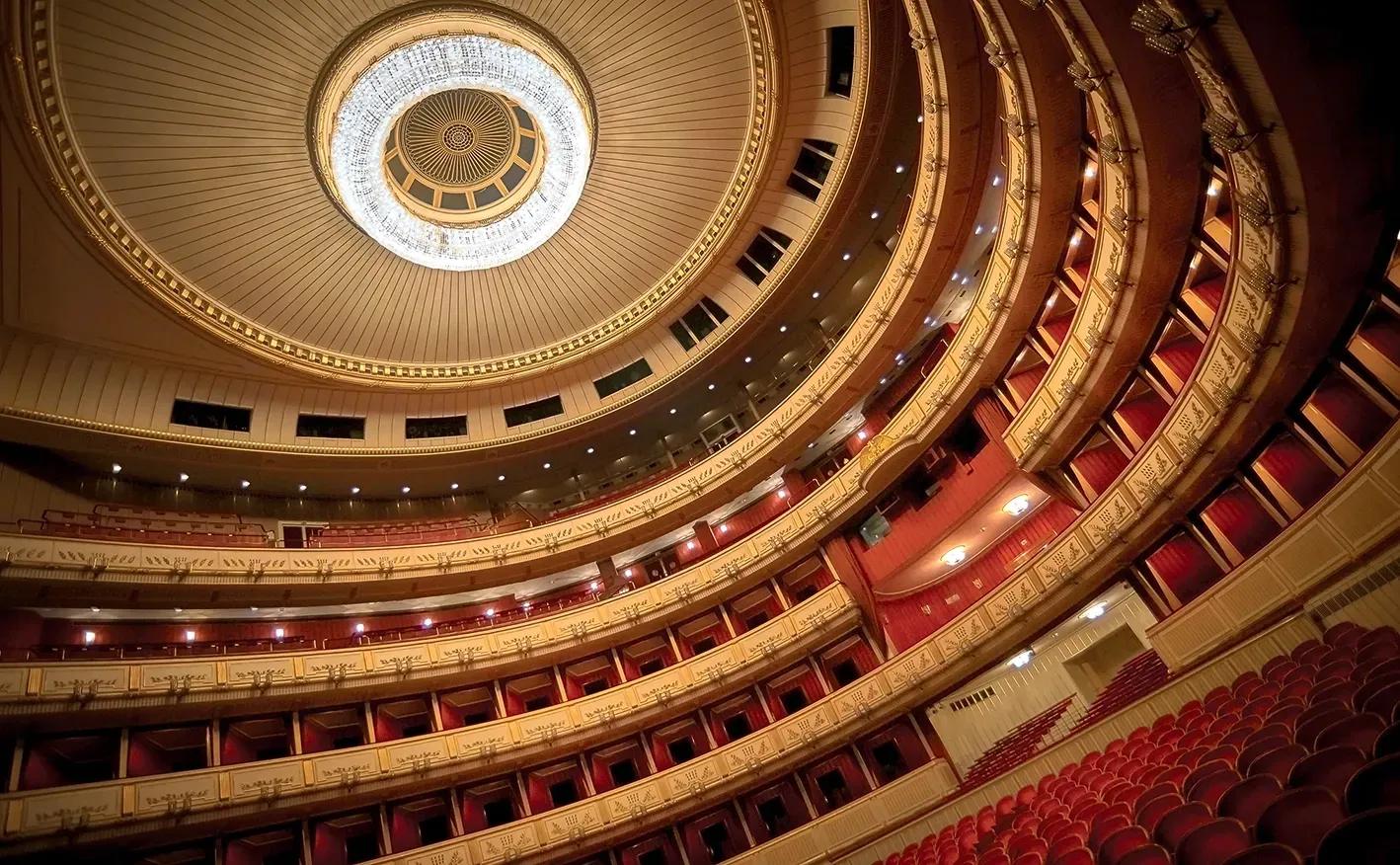 Interior view of a grand opera house. Red seating in tiers ascends to a large circular ceiling with a chandelier.