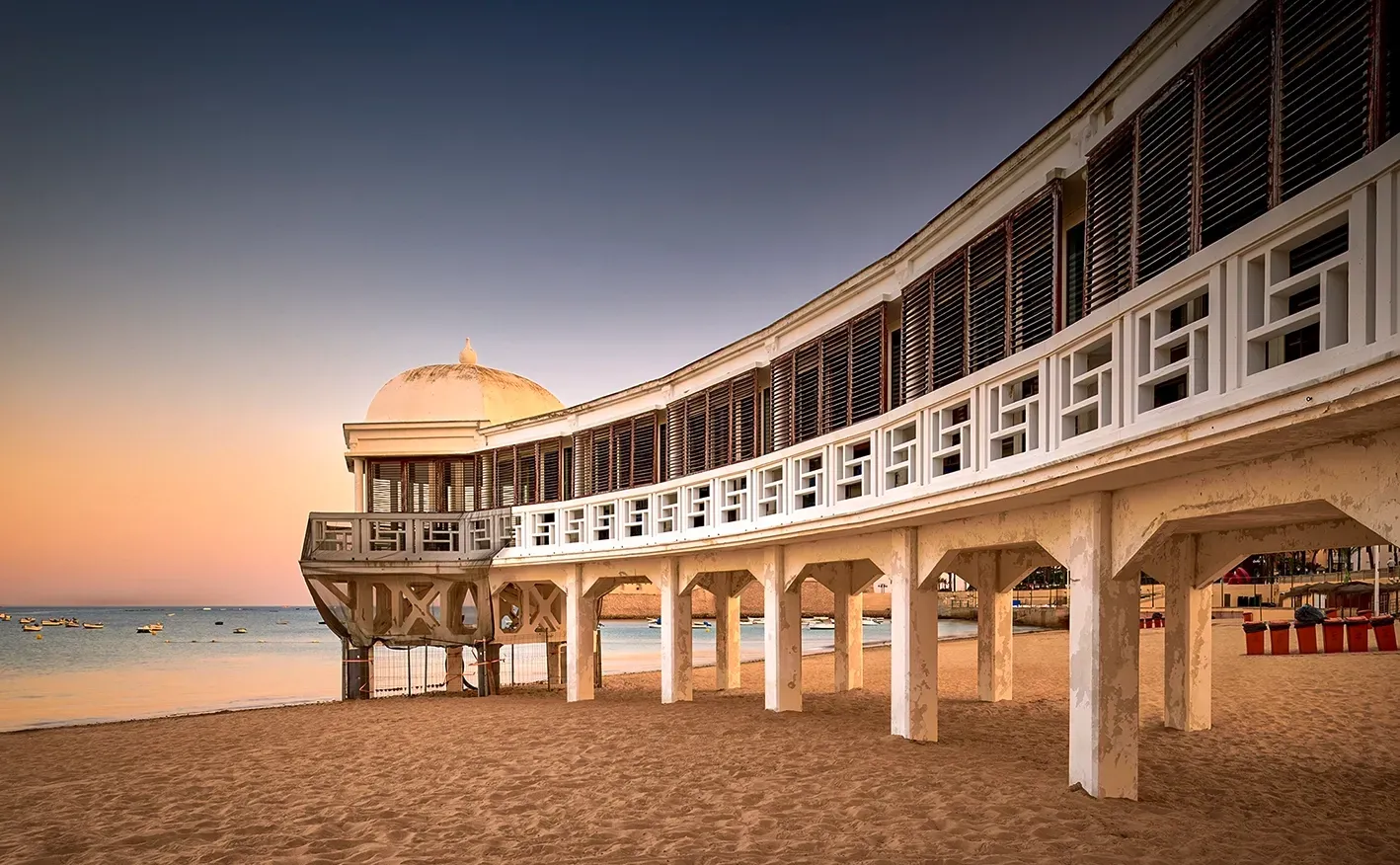Beachfront pavilion on a sandy shore at sunset; white pillars and curved facade.