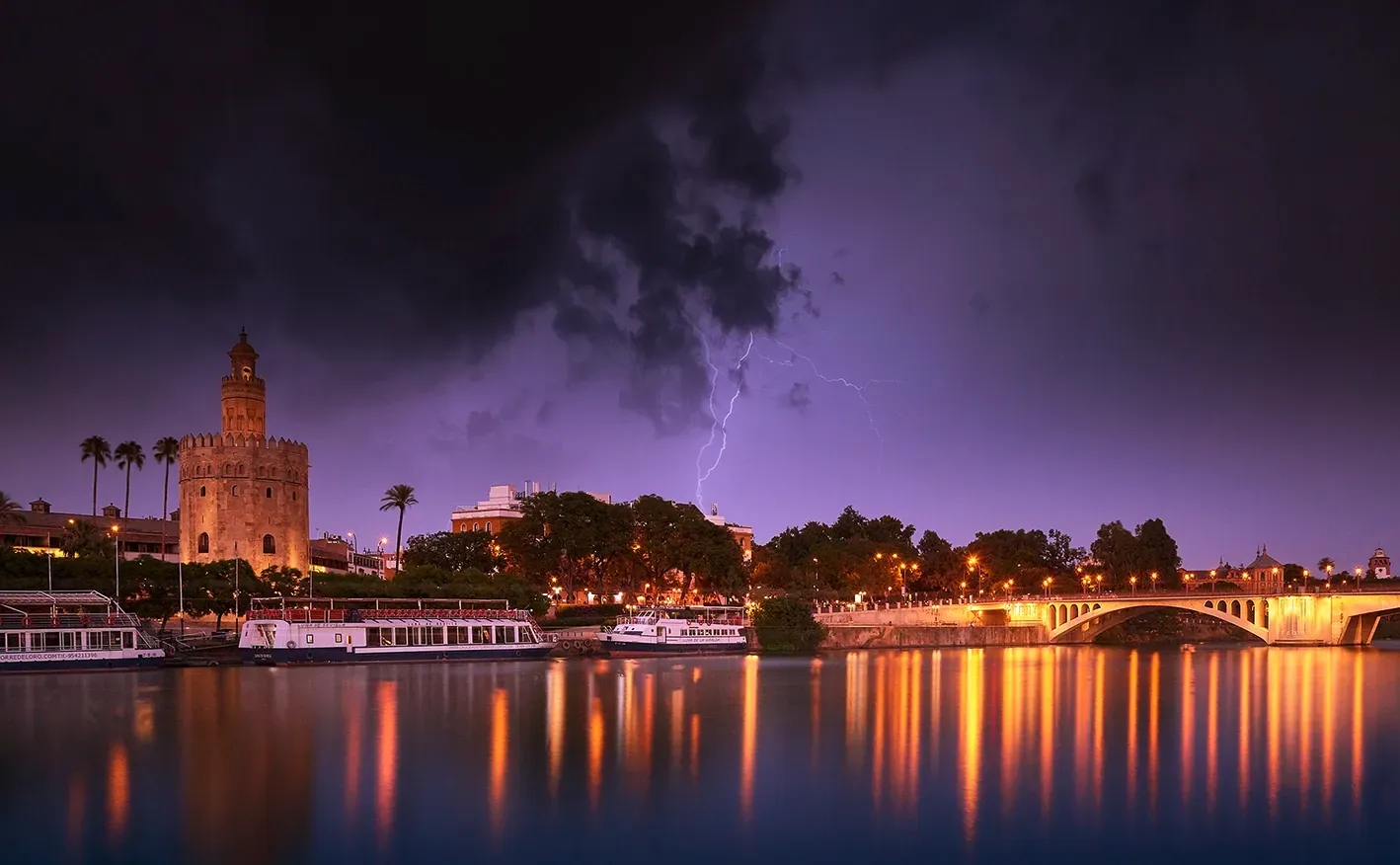 Golden tower and bridge illuminated at night with a lightning strike over a river.