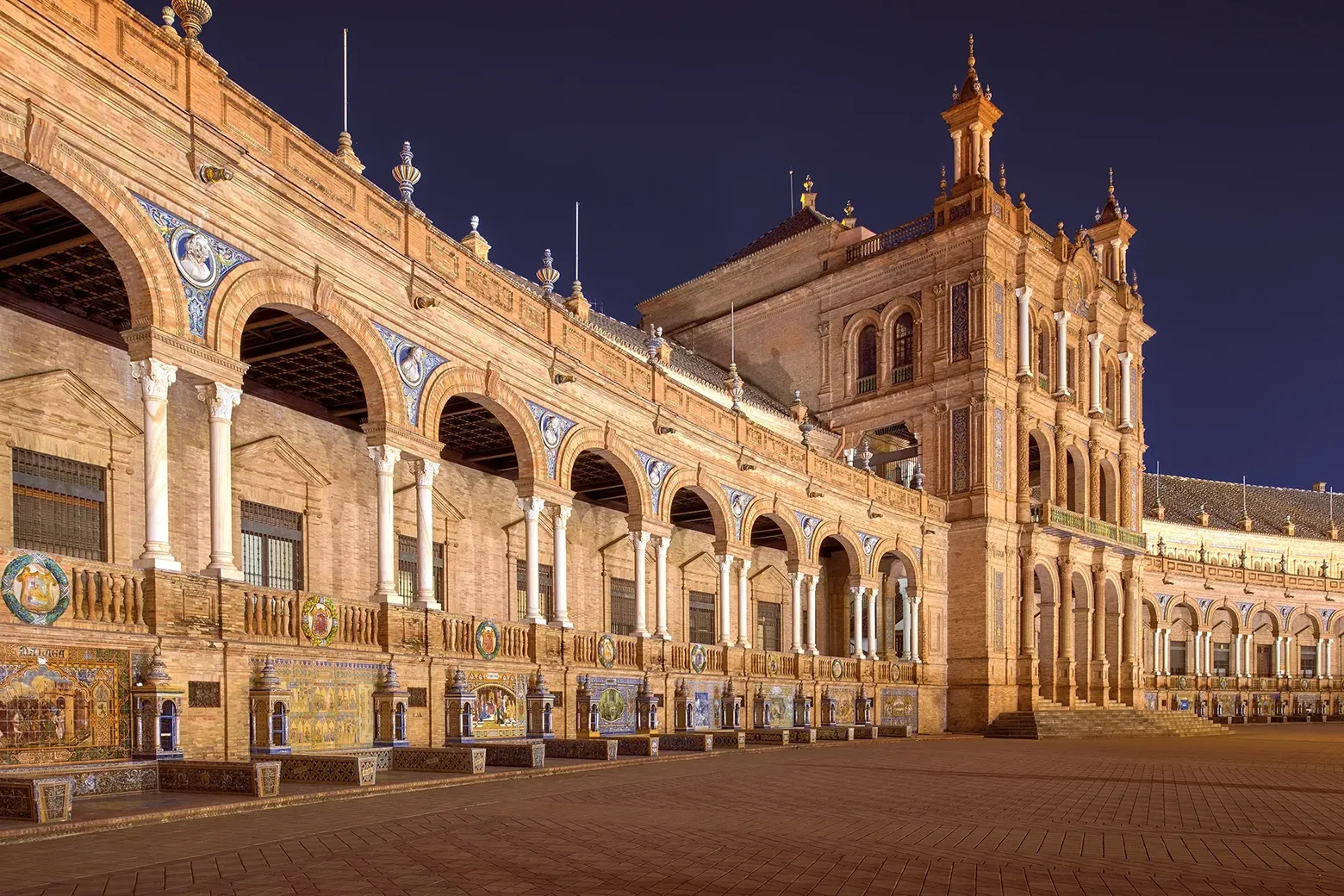 Brick building with arches and tiled murals at Plaza de España, Seville, Spain, lit at night.