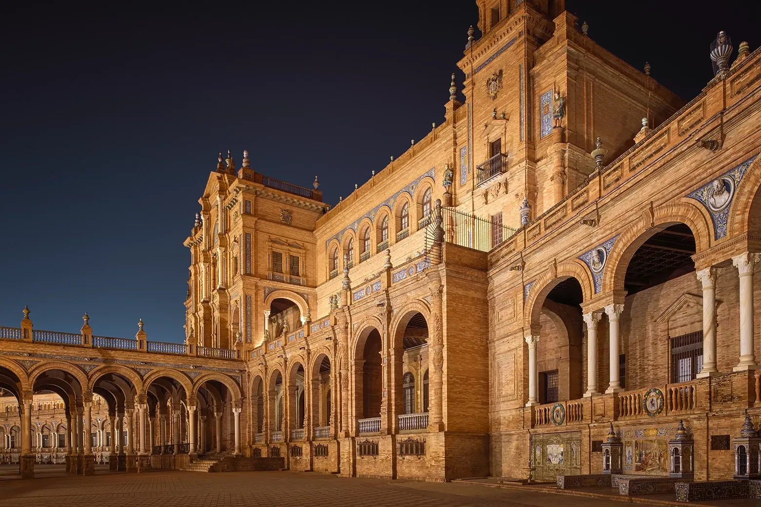 Night view of Plaza de España in Seville, Spain. Illuminated ornate brick building with archways and towers.