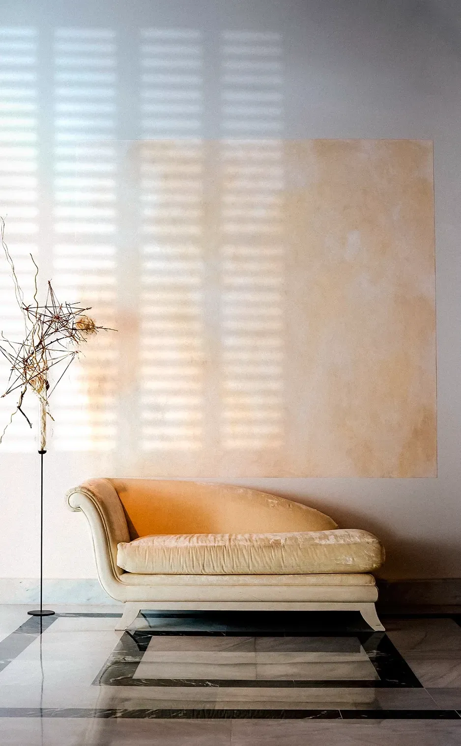Cream-colored chaise lounge next to a tall, decorative branch, set against a wall with sunlight creating a striped effect.