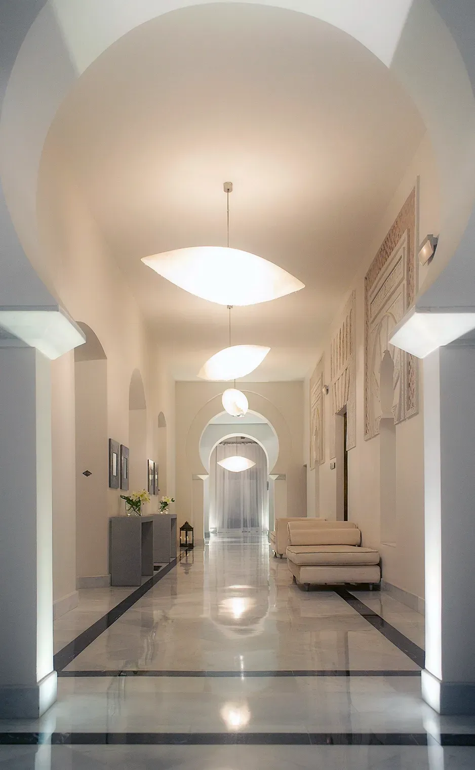Long, white hallway with arched doorways and marble floors. Decorative lights and furniture.