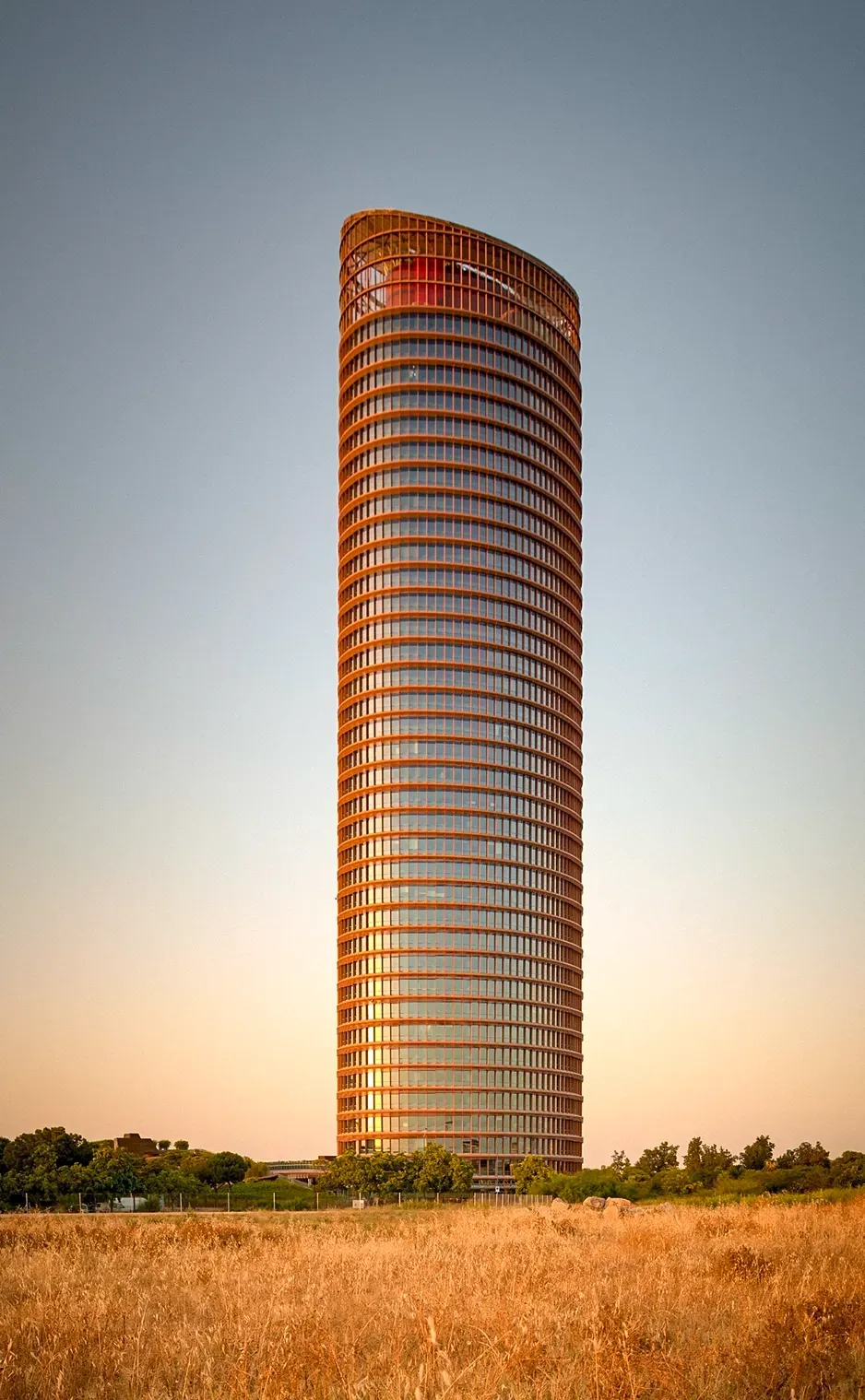 Tall, slender cylindrical skyscraper with curved top, golden glass exterior, set in field, against a blue sky.