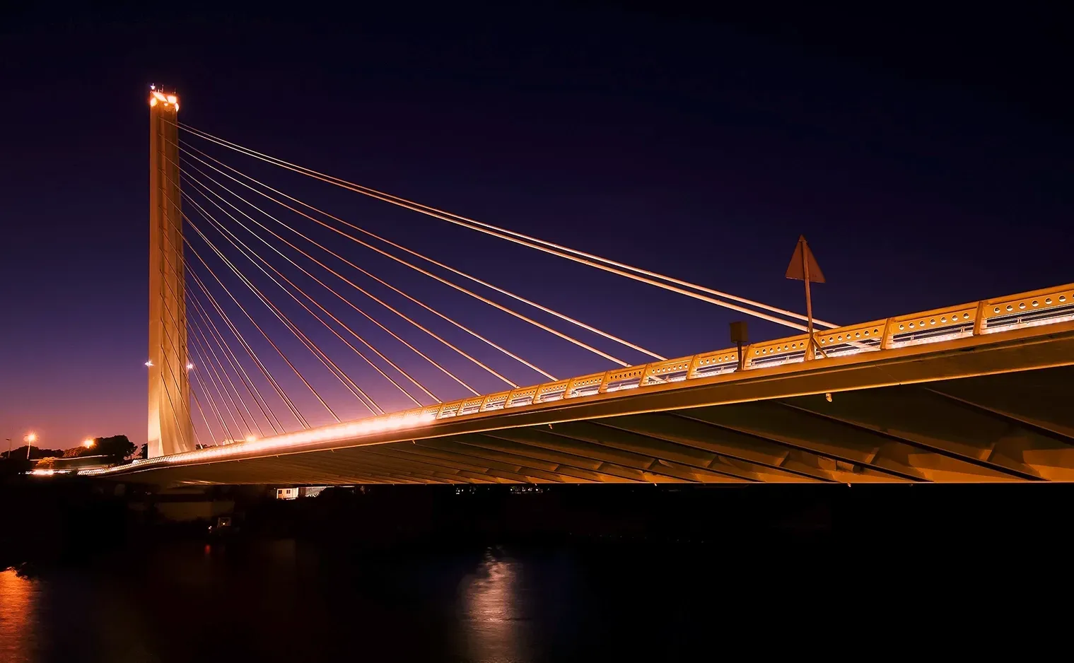 Cable-stayed bridge lit at night. Tall pylon with suspension cables, spanning dark water; lights on the bridge.