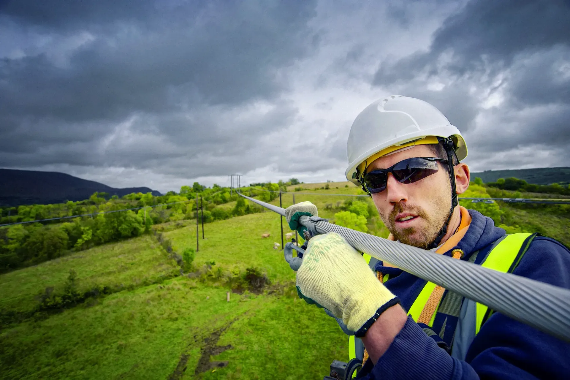 Lineman in safety gear working on a power line, outdoors on a cloudy day.