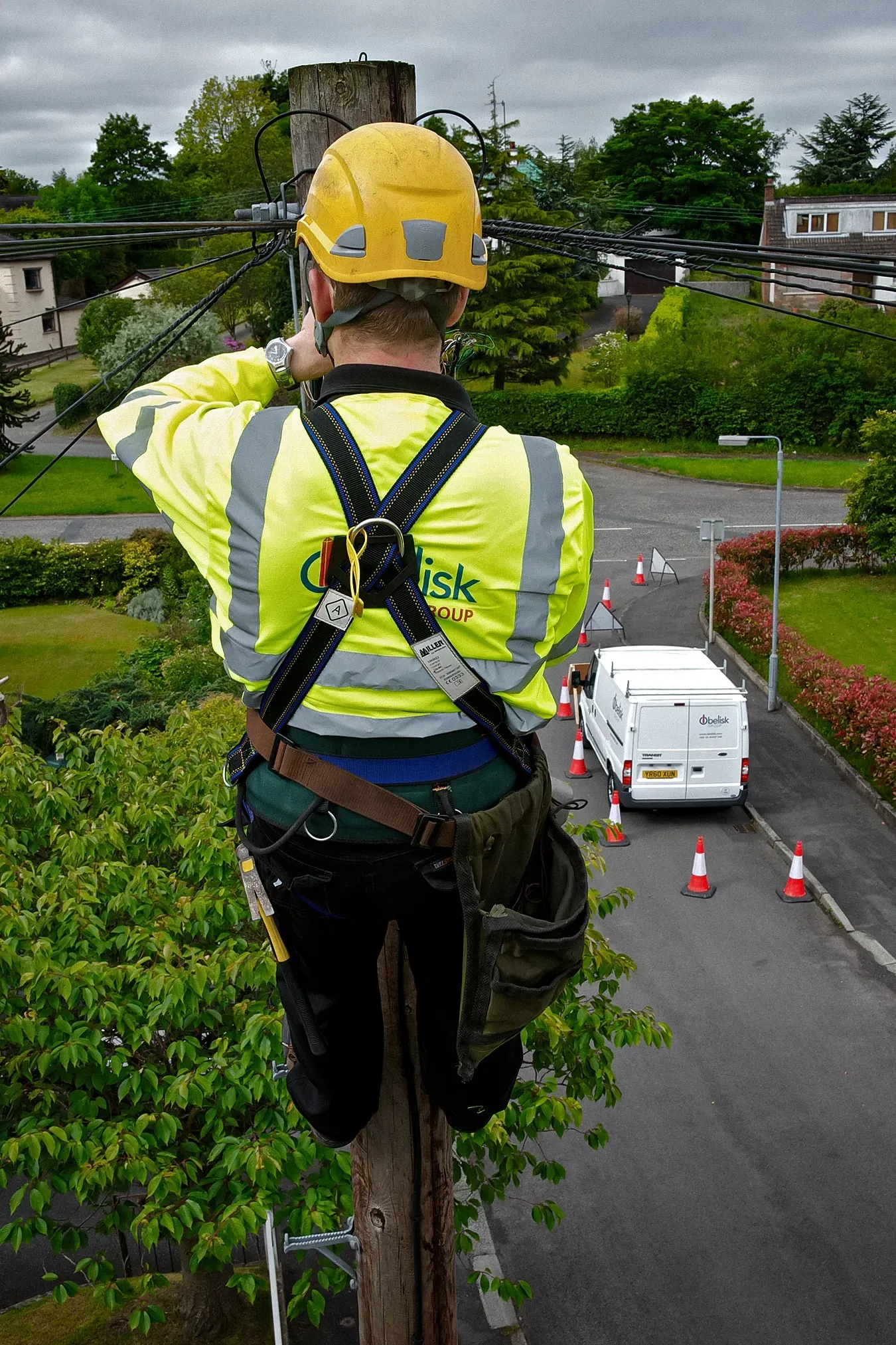 Lineman on a utility pole, wearing a harness and reflective vest, working on overhead wires.