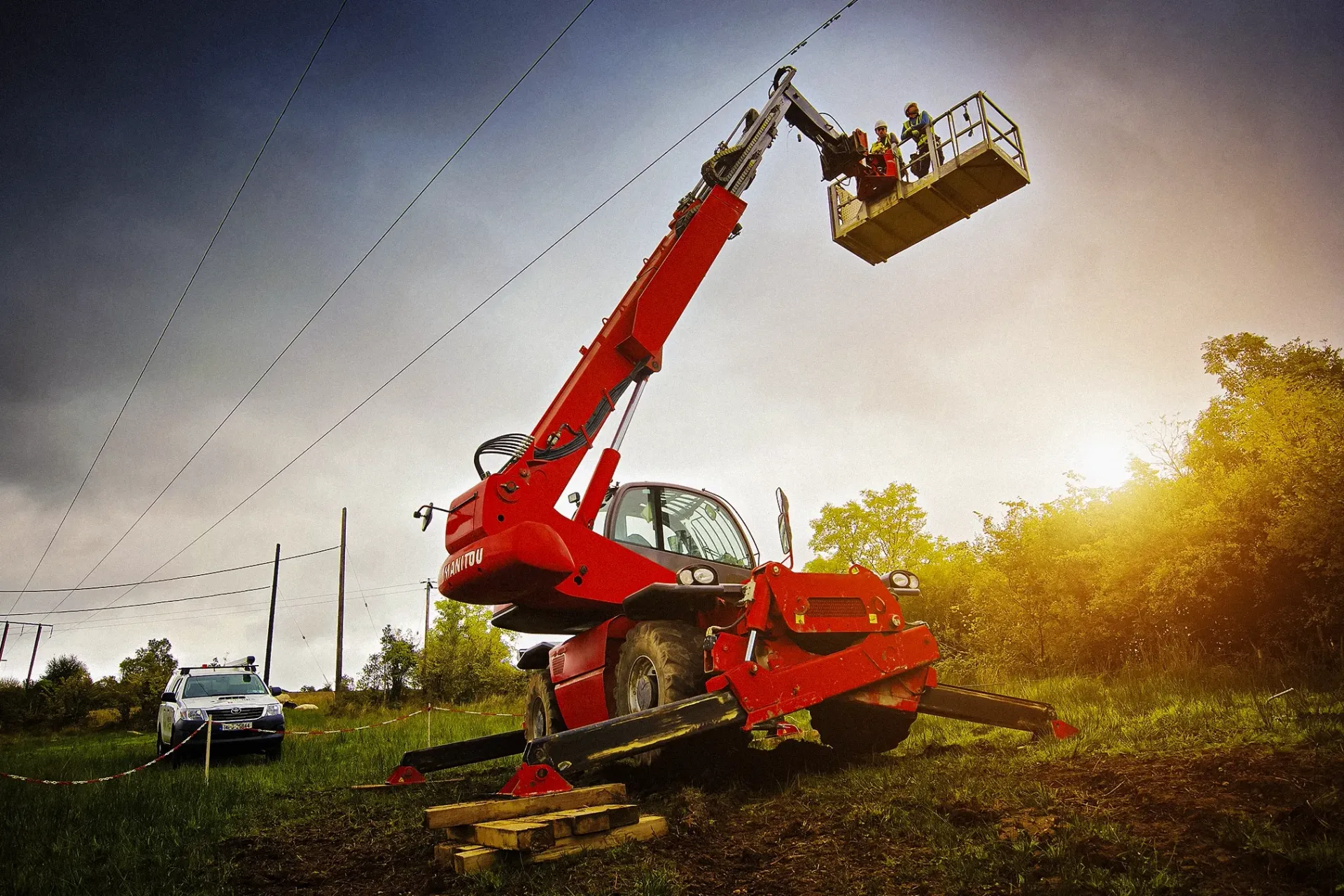 Red telehandler with workers in a basket near power lines. Overcast sky and sunlight.