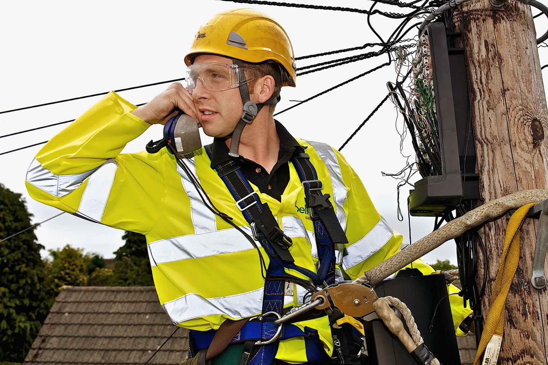 Lineman in yellow safety gear on a utility pole, talking on a radio.