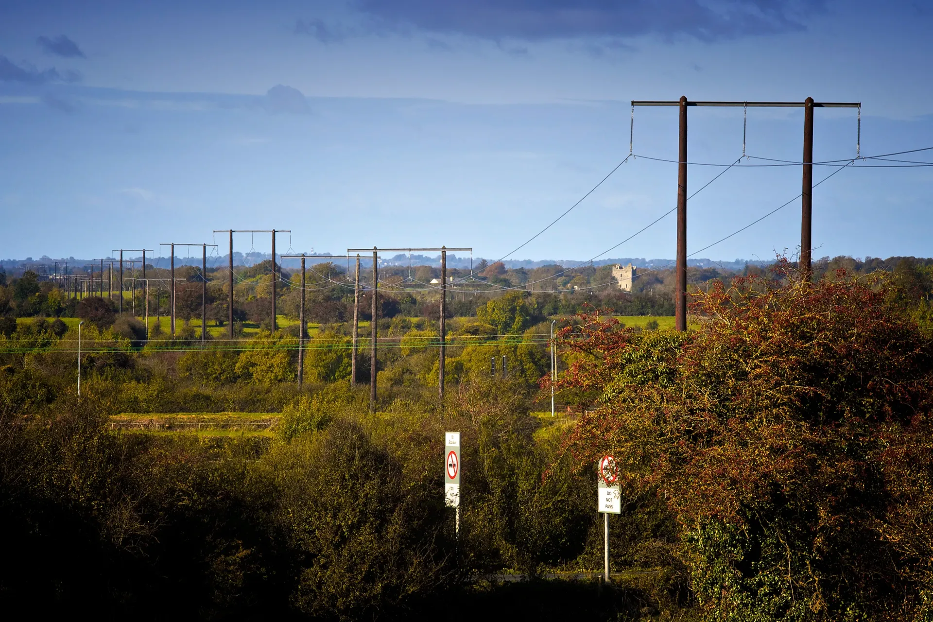 Power lines stretching across a green, shrub-filled landscape under a blue sky.