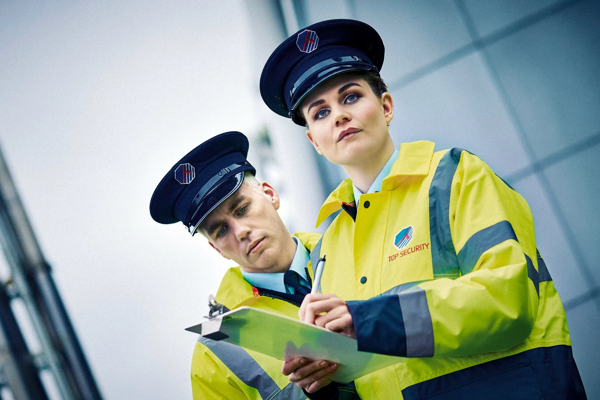 Two uniformed people in yellow jackets and caps, one writing on a clipboard outdoors.