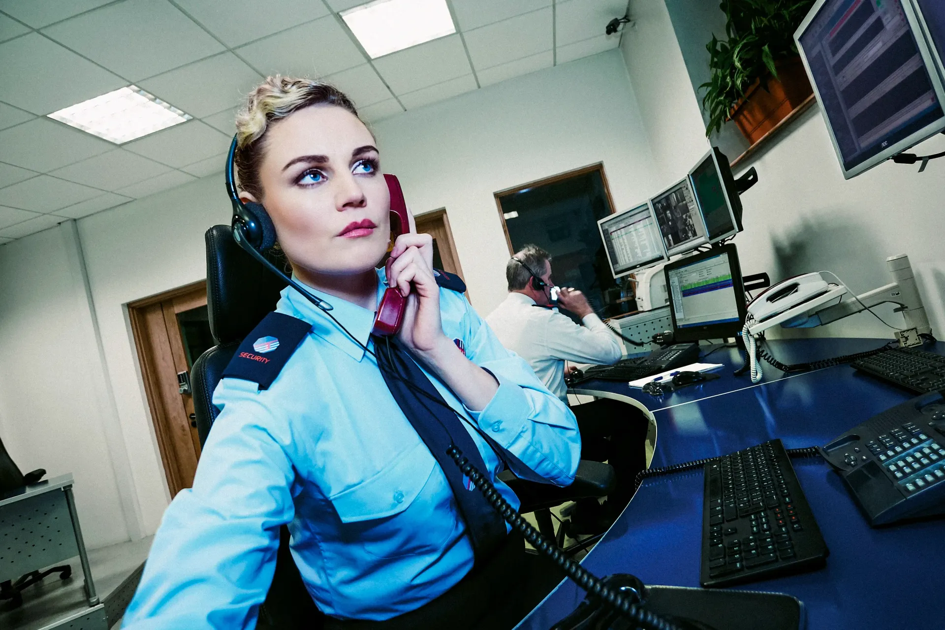 Woman in blue uniform on phone at a control center. A person behind her works at a computer.
