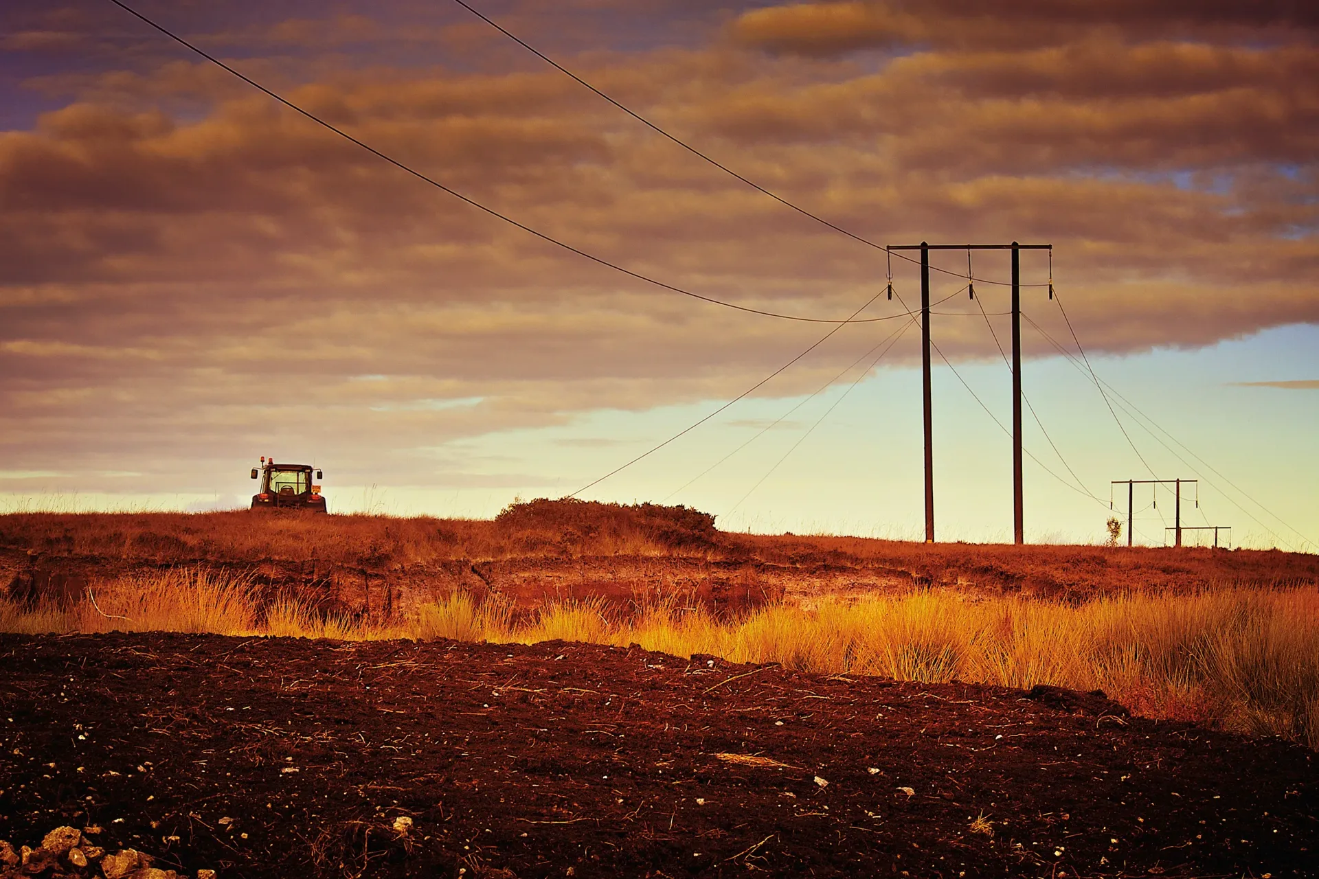 Tractor on a field under power lines at sunset with orange and brown tones.