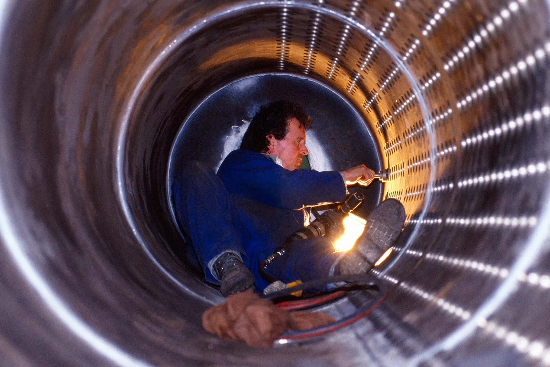 A worker inside a large metal pipe, welding. He's wearing blue coveralls, with his legs braced.