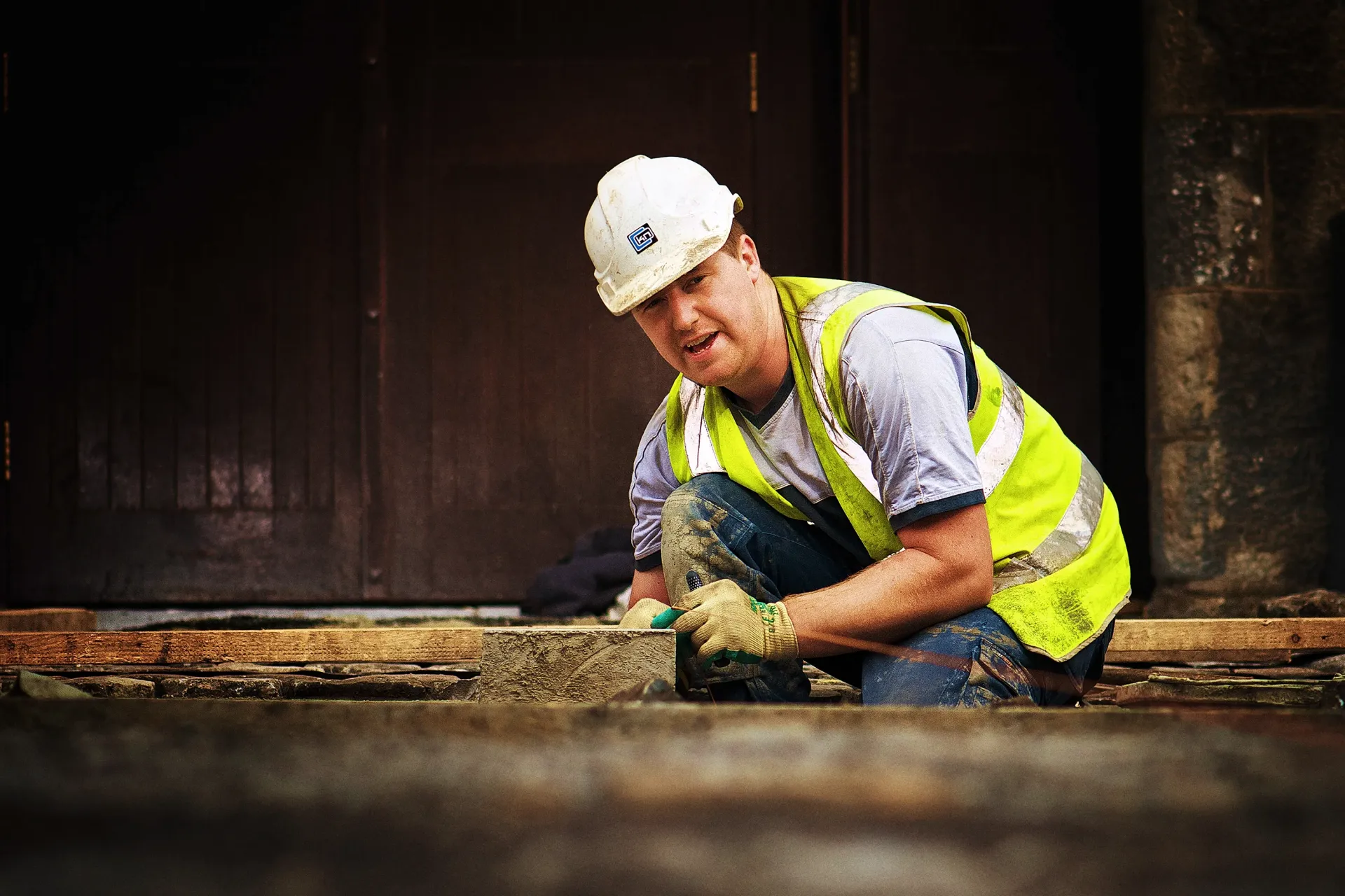 Construction worker in white hard hat and yellow vest kneels to work on a building's foundation.