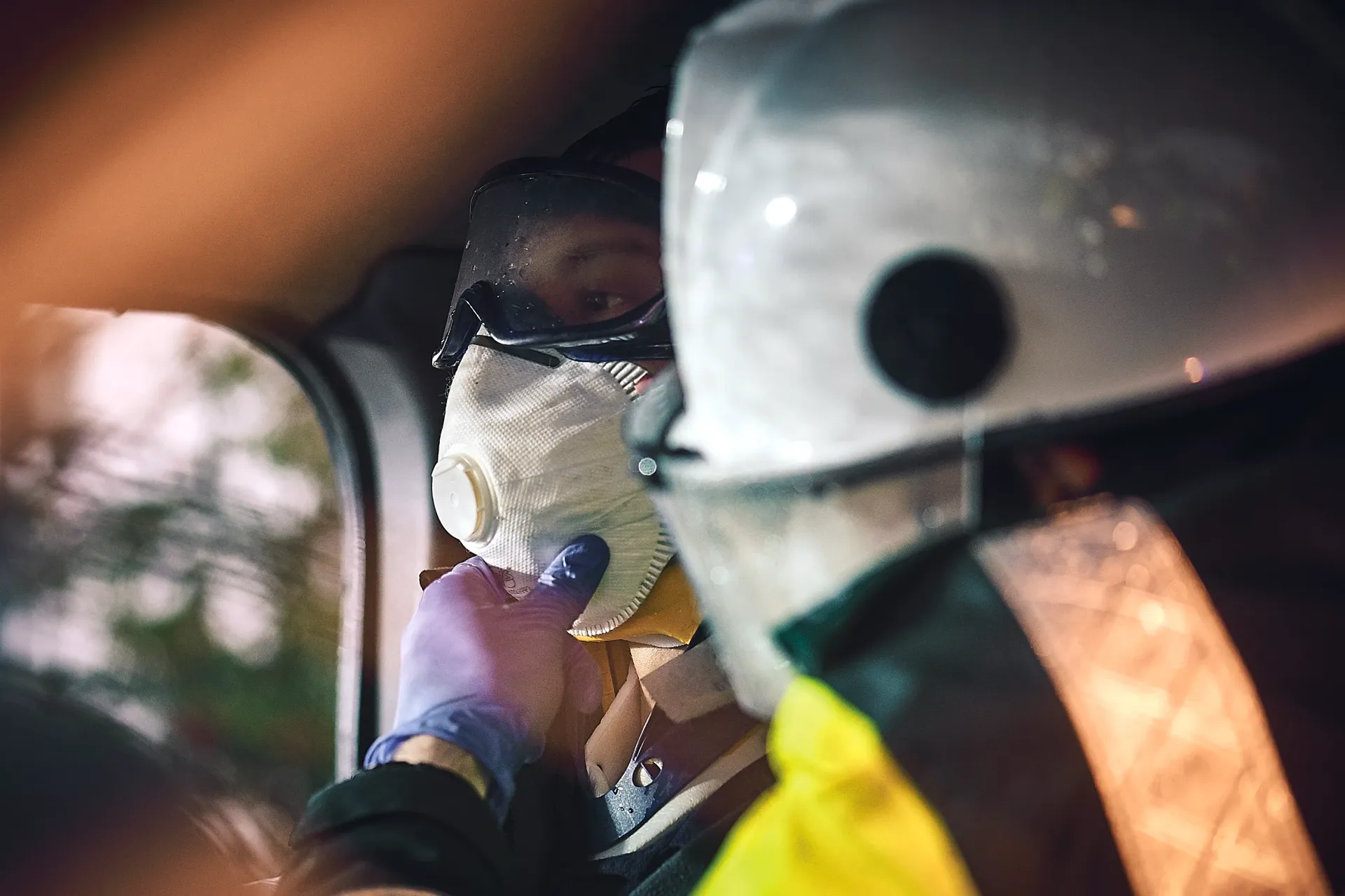 Emergency personnel treating a person wearing a face mask inside a vehicle; low light.