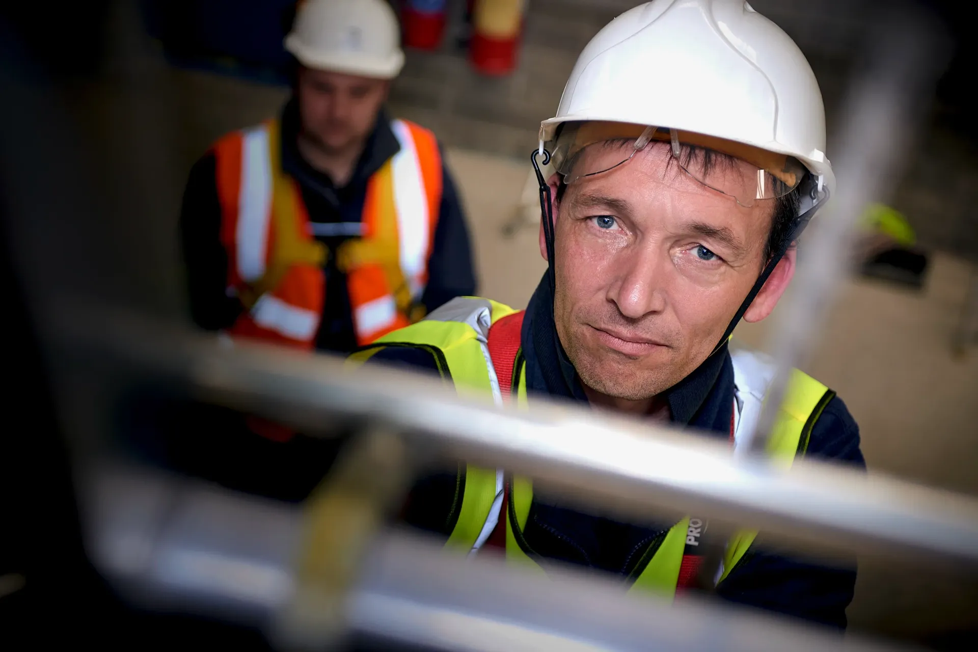 Two construction workers in hard hats and vests on a metal structure; one looks up.