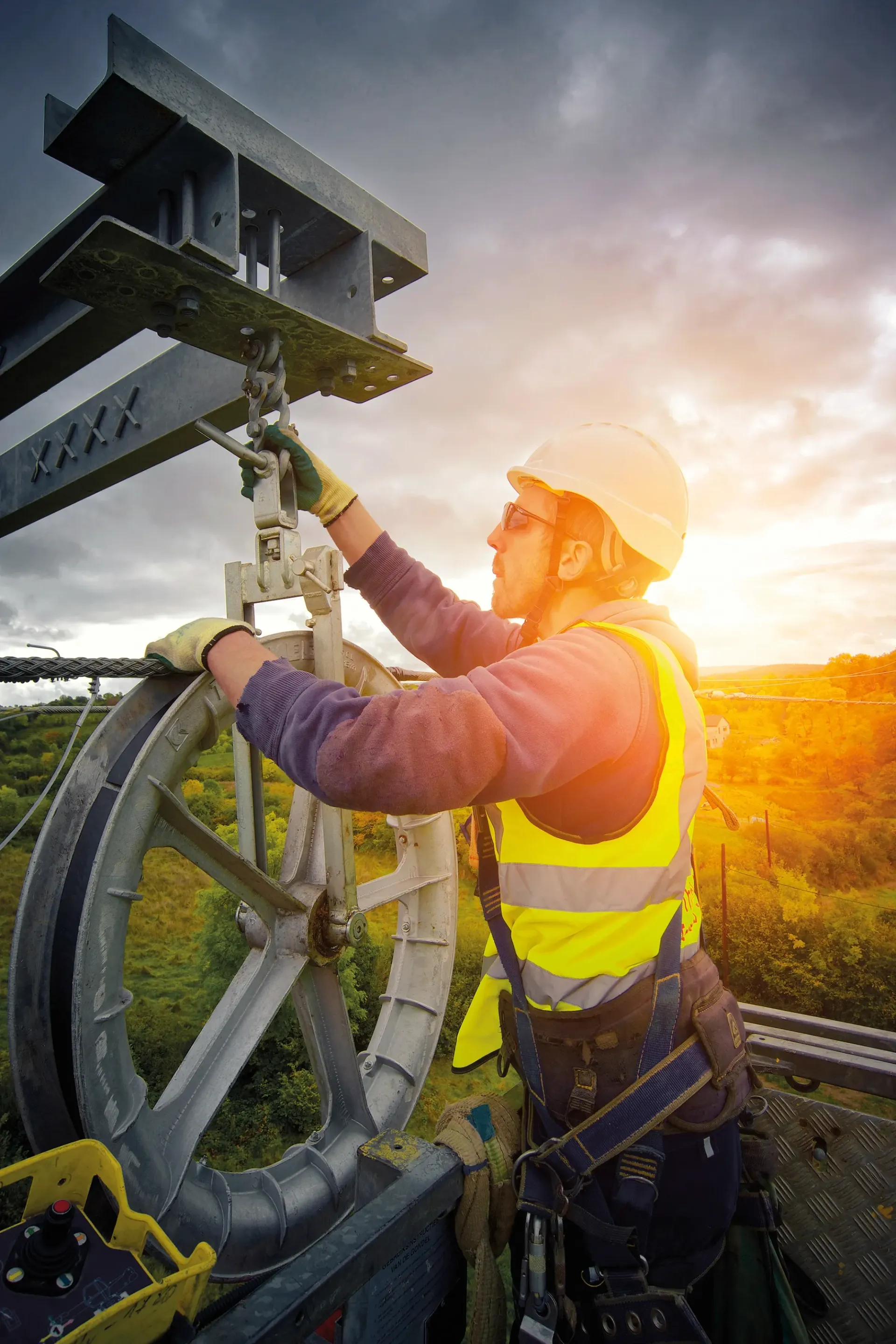 Worker in safety gear adjusting a large pulley system outdoors with a sunset in the background.