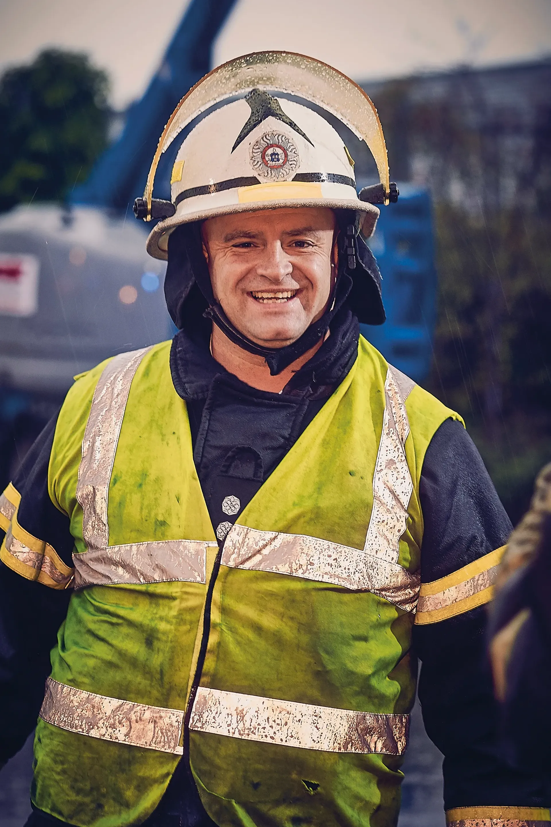 Firefighter wearing helmet and high-visibility vest smiles in front of a blue lift.